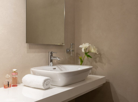 Ein modernes Waschbecken von SHK Technik mit einem verchromten Wasserhahn, ein weißes Handtuch auf Rollen, zwei kleine Flaschen und eine Vase mit weißen Blumen stehen auf einer schlichten weißen Arbeitsplatte unter einem quadratischen Spiegel an einer beigen Wand.