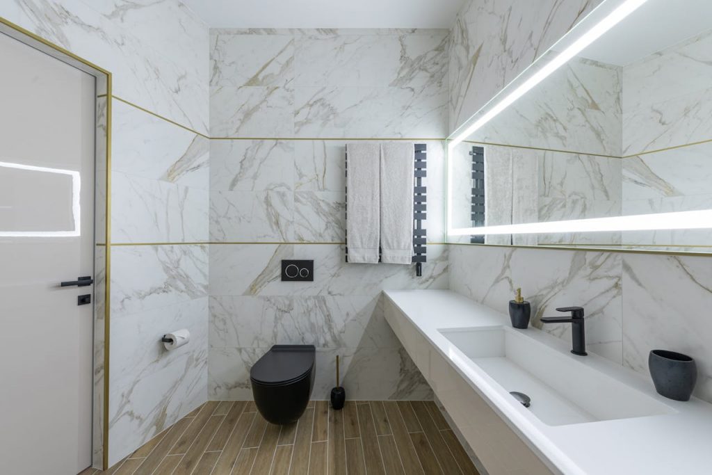 Modernes Badezimmer mit marmorierten Wänden, Holzboden, schwarzem WC und Armaturen, langem weißen Waschbecken, zwei Handtüchern und einem beleuchteten Spiegel mit hochwertigen SHK-Technik-Elementen